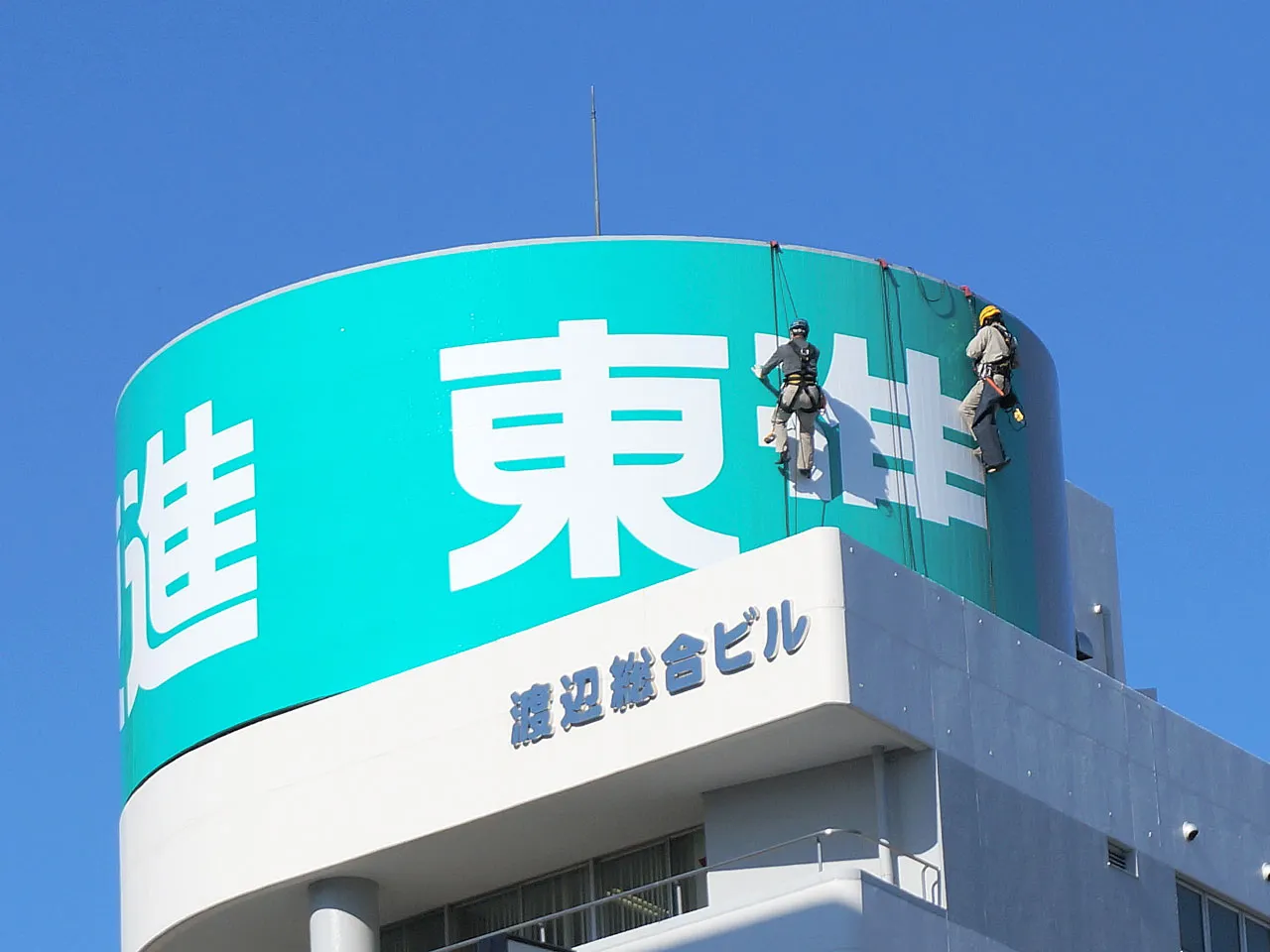 ロープアクセスでシート貼りする塔屋看板